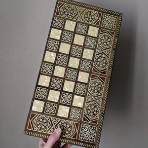 Può includere: Un tabellone di gioco rettangolare in legno con un design intarsiato. Il tabellone presenta un motivo a scacchiera con quadrati chiari e motivi geometrici nei toni del marrone, rosso e bianco sui bordi. Il tabellone è tenuto da una mano.