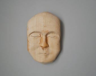 Pode incluir: Uma escultura de madeira de um rosto com os olhos fechados. A escultura é esculpida em um estilo realista e tem uma cor marrom claro.