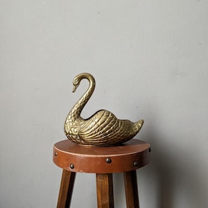 May include: A vintage brass swan figurine with detailed feather textures. The swan is perched on a small, round, brown wooden stool with dark wooden legs. The stool has metal studs around the edge of the seat.