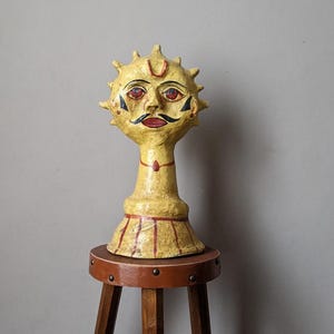 Puede incluir: Una escultura de cerámica amarilla de una cara con ojos rojos, un bigote negro y un punto rojo en la frente. La escultura está sobre un taburete de madera.
