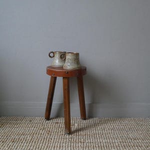 May include: Two ceramic mugs with a brown and white speckled glaze sit on a wooden stool with three legs. The stool has a brown leather top with decorative brass studs.