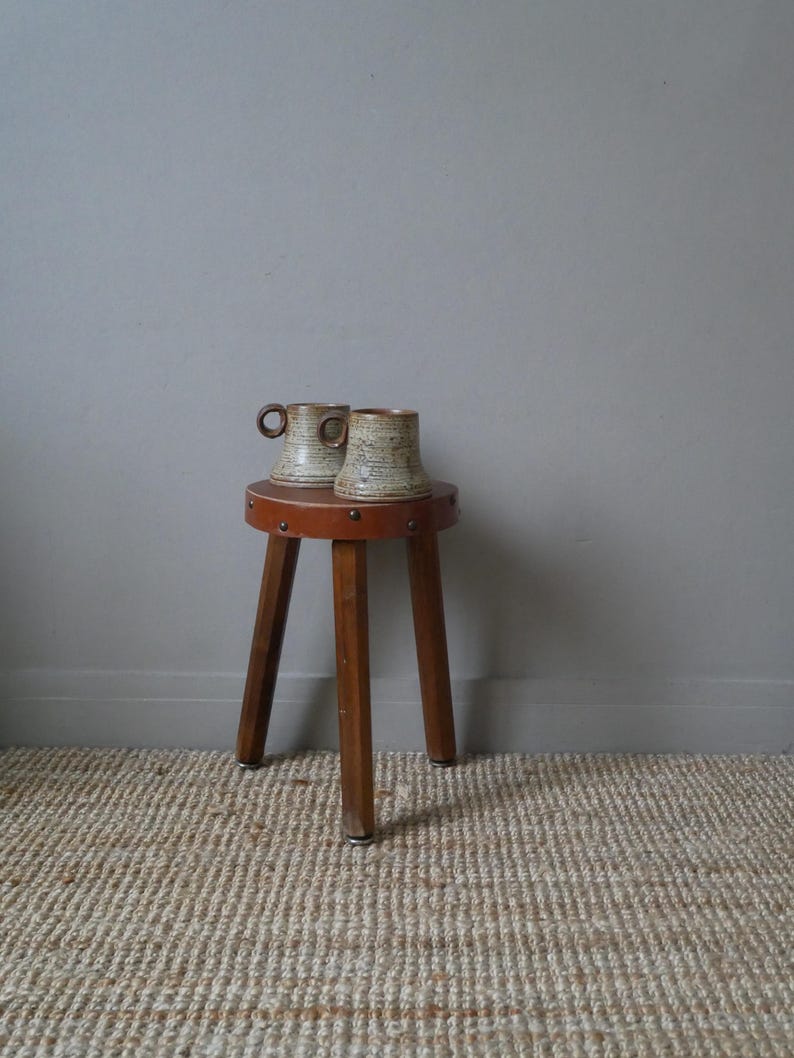 May include: Two ceramic mugs with a brown and white speckled glaze sit on a wooden stool with three legs. The stool has a brown leather top with decorative brass studs.
