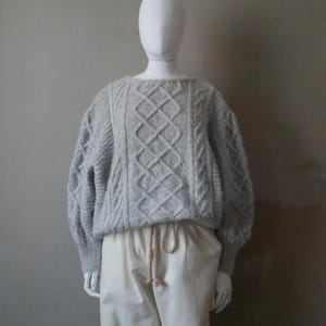 Peut inclure: Un pull gris en tricot torsadé à la coupe ample et aux manches longues. Le pull présente un motif en losanges.