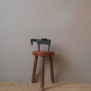 Peut inclure: Une sculpture de vache en métal noir est posée sur un tabouret en cuir marron à trois pieds en bois. Le tabouret est sur un tapis tissé.
