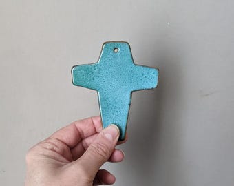 Könnte beinhalten: Ein türkisfarbenes Keramik-Kreuz mit strukturierter Oberfläche und dunklem braunen Rand. Das Kreuz hat oben ein Loch zum Aufhängen. Das Kreuz wird in der Hand einer Person vor einem neutralen Hintergrund gehalten.