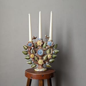 Könnte beinhalten: Ein Kerzenhalter aus Keramik in Form eines Baumes mit braunen, grünen, gelben und blauen Blumen. Drei weiße Kerzen stehen im Halter. Der Halter steht auf einem Holzhocker.