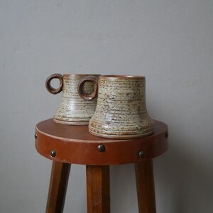 May include: Two brown and white ceramic mugs with a textured finish sit on a brown wooden stool with a leather top.