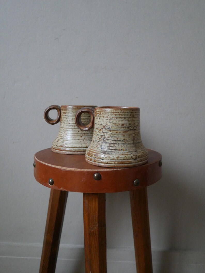 May include: Two brown and white ceramic mugs with a textured finish sit on a brown wooden stool with a leather top.