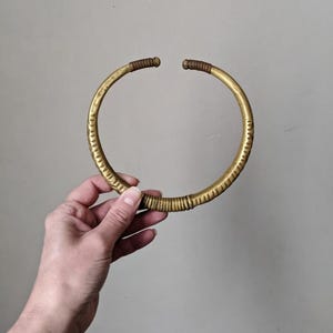 Op de afbeelding: Een goudkleurige, open ketting met een getextureerd oppervlak. De ketting heeft een cirkelvorm met gewikkelde details nabij de uiteinden. De uiteinden zijn afgedekt met een bruin materiaal. Het sieraad wordt door een hand vastgehouden.