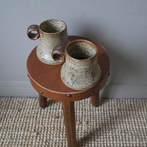 May include: Two brown and white speckled ceramic mugs with wooden handles sitting on a brown leather stool with wooden legs.