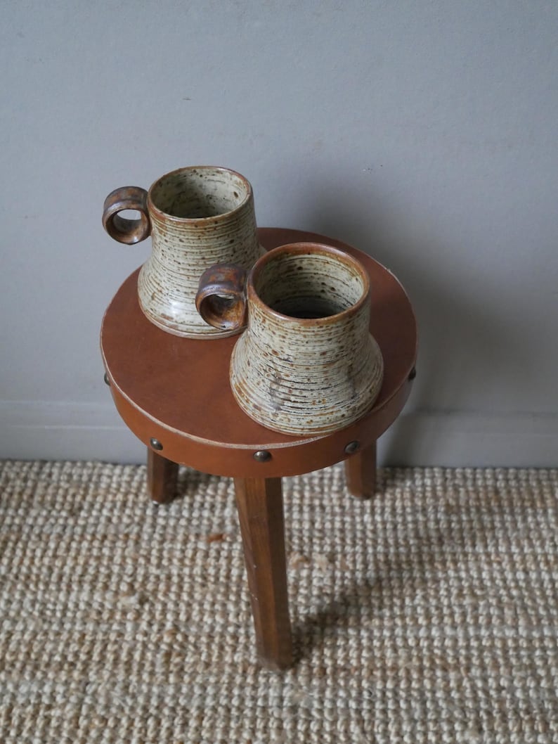 May include: Two brown and white speckled ceramic mugs with wooden handles sitting on a brown leather stool with wooden legs.