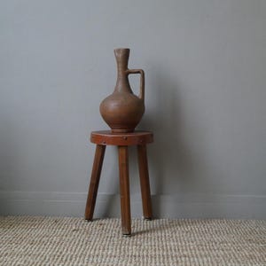 Könnte beinhalten: Eine braune Keramikvase mit Henkel steht auf einem Holzhocker mit braunem Ledersitz. Der Hocker hat drei Beine und steht auf einem gewebten Teppich.