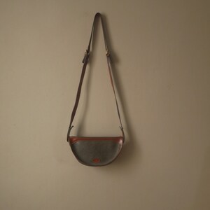 Könnte beinhalten: Eine braune Lederschultertasche mit einer grauen Stoffklappe und einem braunen Lederbesatz. Die Tasche hängt an einer hellbeigen Wand.