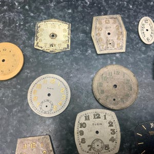 May include: A collection of vintage watch faces in various shapes and sizes. The faces are primarily off-white with black or gold numerals and hands. Some faces have the brand name "Elgin" printed on them.