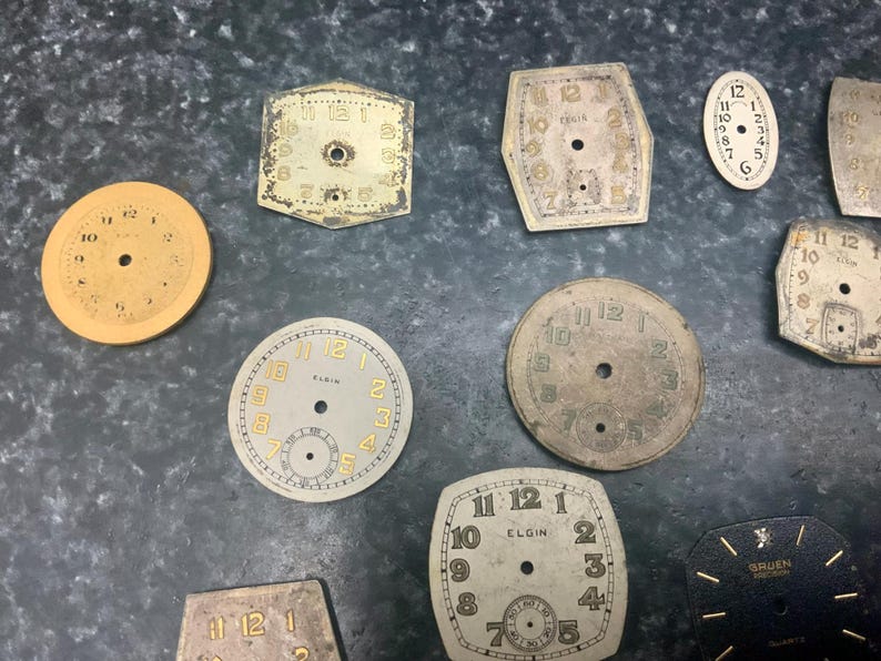 May include: A collection of vintage watch faces in various shapes and sizes. The faces are primarily off-white with black or gold numerals and hands. Some faces have the brand name "Elgin" printed on them.