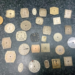 May include: A collection of vintage watch faces in various shapes, including round, square, and oval. The faces are primarily cream and gold, with black numerals and hands. Some show signs of age and wear.