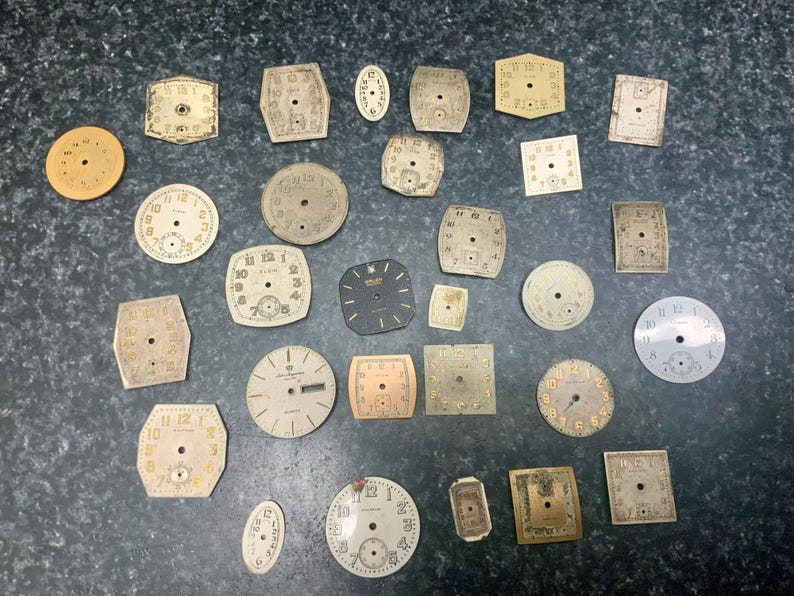 May include: A collection of vintage watch faces in various shapes, including round, square, and oval. The faces are primarily cream and gold, with black numerals and hands. Some show signs of age and wear.