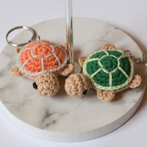 Könnte beinhalten: Zwei gehäkelte Schildkröten-Schlüsselanhänger, einer mit einer orangefarbenen Schale und der andere mit einer grünen Schale. Beide Schildkröten haben beigefarbene Körper und schwarze Augen.