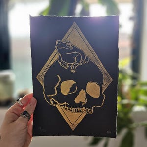 May include: A black and gold print of a skull with a frog sitting on top. The skull and frog are outlined in gold and the background is black. The print is signed and numbered "19/20" and "8/11".