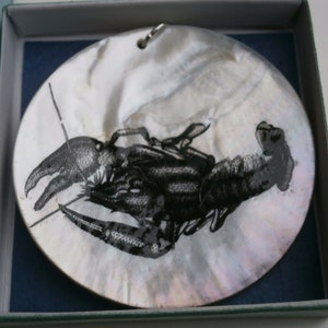 May include: A round, iridescent, mother-of-pearl ornament featuring a black and white image of a crayfish.