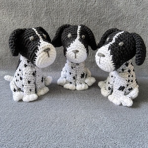 May include: Three handmade crochet dog figurines. Each dog is white with black spots and black ears. The dogs are sitting upright with their paws in front. The background is a solid gray.