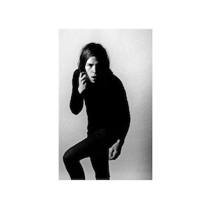 May include: A black and white photo of a person with long hair wearing a black turtleneck shirt and black pants. The person is standing with one leg bent and their hand near their face.