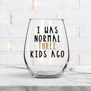 May include: A clear glass wine glass with the text "I was normal three kids ago" printed in black and gold on the side.