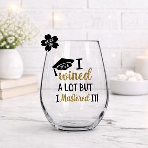 May include: A clear stemless wine glass with the text "I wined a lot but I Mastered it!" in gold glitter lettering. A black graduation cap graphic with the year 2026 is above the text. A small black flower with the text "Name can be added" is on the rim.