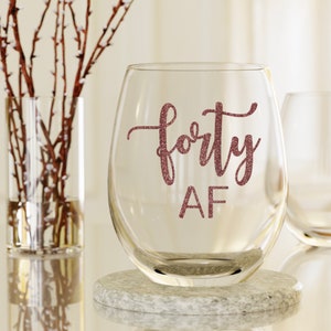 May include: A clear stemless wine glass with the words "forty AF" in glittery pink script. The glass sits on a small, round, gray stone coaster. A second glass and a vase with branches are in the background.