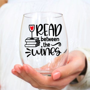 May include: A clear glass wine glass with a black and white design that says "Read between the wines". The design also includes a red heart and a stack of books.