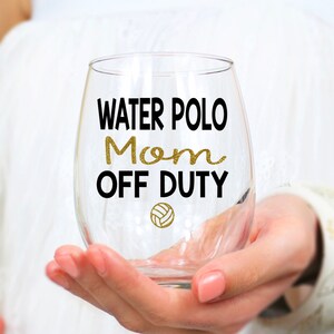 May include: A clear stemless wine glass with the words "WATER POLO Mom OFF DUTY" in black and gold lettering. A gold volleyball graphic is below the text. The glass is held by a person.