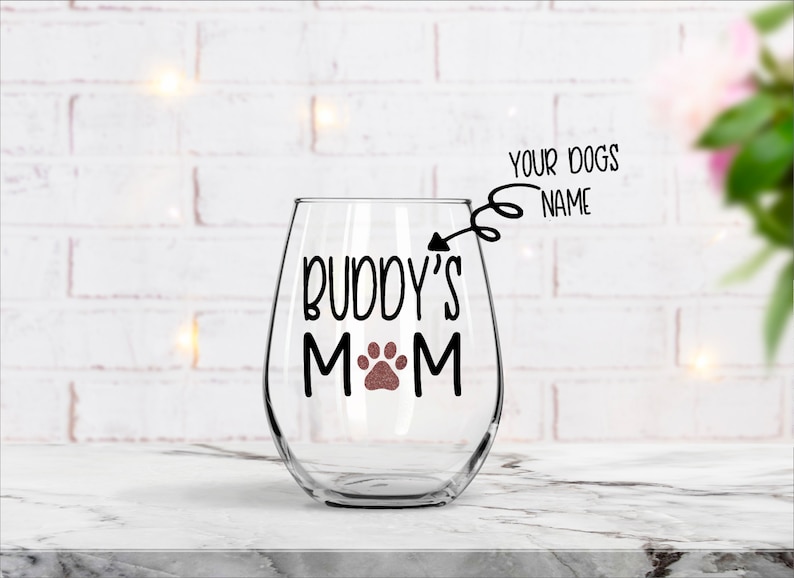 May include: A clear glass wine glass with a black and brown paw print design and the text "Buddy's Mom". The text "Your Dogs Name" is written above the glass.