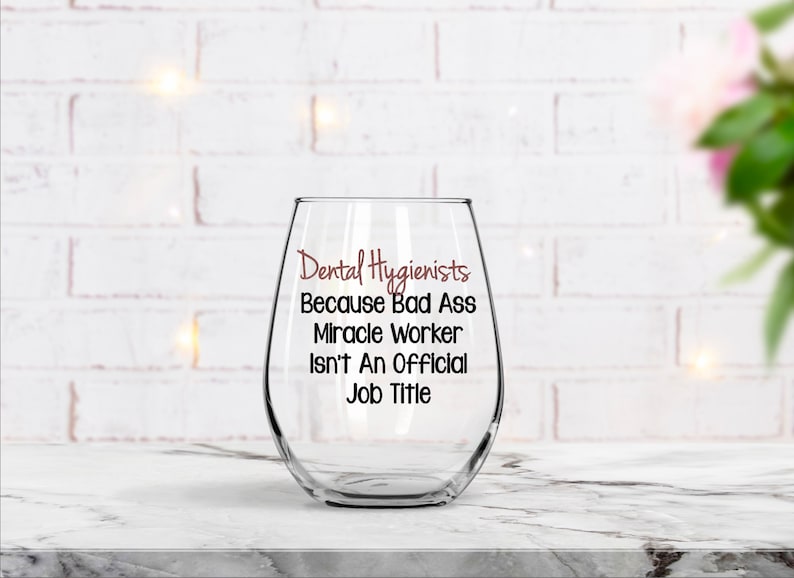 May include: A clear glass wine glass with the text "Dental Hygienists Because Bad Ass Miracle Worker Isn't An Official Job Title" printed on it.