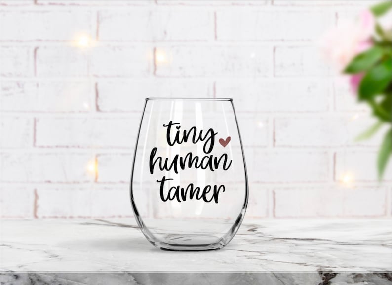K&ouml;nnte beinhalten: Ein klares Weinglas aus Glas mit dem Text "tiny human tamer" in schwarzer Schrift und einem kleinen roten Herzen.