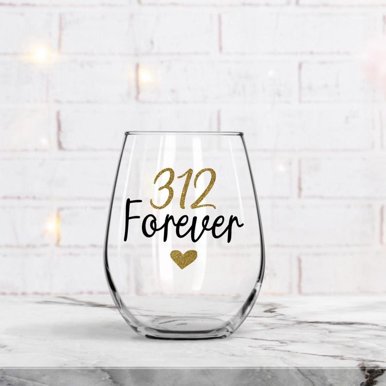 Op de afbeelding: Een helder glazen wijnglas met een gouden glitterhart en de tekst "312 Forever" in zwarte letters.