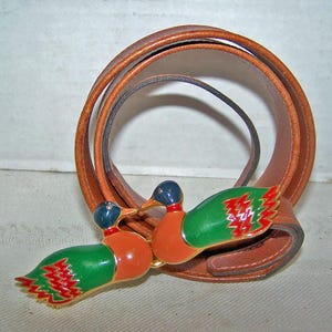 May include: A brown leather belt with a decorative buckle featuring two colorful duck figures. The ducks have green wings, orange bodies, and blue heads. The belt is a light brown color.
