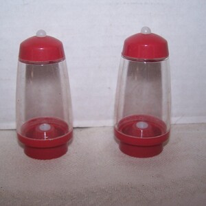 May include: Two red and clear plastic salt and pepper shakers. Each shaker has a red base and lid, with a clear, conical body. A small white button is on the top of each lid. The shakers are designed to sit upright.