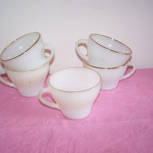 May include: A collection of six white teacups with gold rims and handles. The cups are stacked and arranged on a pink surface. The cups have a classic design, suitable for serving tea or coffee.