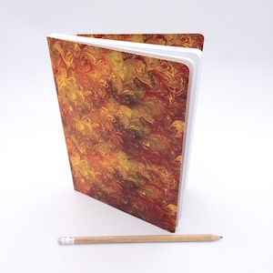 Pode incluir: Um caderno fechado com uma capa marmorizada em tons de vermelho, laranja e amarelo. O caderno tem páginas brancas e está sobre uma superfície branca.