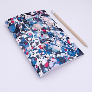 May include: A notebook with a marbled cover in shades of blue, black, white, pink, and red. The cover has a swirling pattern of circles and dots. A wooden pencil is lying next to the notebook.