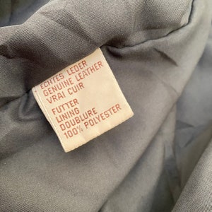 K&ouml;nnte beinhalten: Ein braunes Leder-Etikett mit wei&szlig;em Text, der "Echtes Leder Genuine Leather Vrai Cuir Futter Lining Doublure 100% Polyester" lautet.
