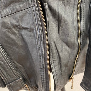 K&ouml;nnte beinhalten: Schwarze Lederjacke mit Rei&szlig;verschluss und G&uuml;rteldetail. Die Jacke hat eine weiche, strukturierte Oberfl&auml;che und ein klassisches Design.