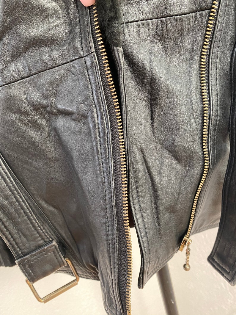 K&ouml;nnte beinhalten: Schwarze Lederjacke mit Rei&szlig;verschluss und G&uuml;rteldetail. Die Jacke hat eine weiche, strukturierte Oberfl&auml;che und ein klassisches Design.