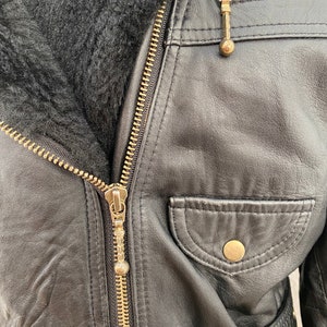 K&ouml;nnte beinhalten: Schwarze Lederjacke mit Rei&szlig;verschluss und Pelzkragen. Die Jacke hat eine Tasche auf der Vorderseite und einen G&uuml;rtel.