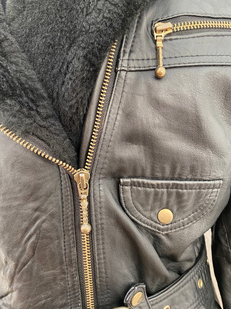 K&ouml;nnte beinhalten: Schwarze Lederjacke mit Rei&szlig;verschluss und Pelzkragen. Die Jacke hat eine Tasche auf der Vorderseite und einen G&uuml;rtel.
