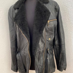 K&ouml;nnte beinhalten: Eine schwarze Lederjacke mit einem Lammfellkragen und einem G&uuml;rtel. Die Jacke hat einen Rei&szlig;verschluss und mehrere Taschen.