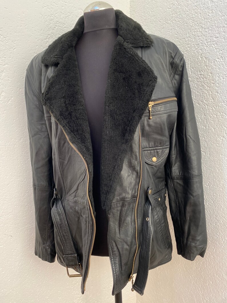 K&ouml;nnte beinhalten: Eine schwarze Lederjacke mit einem Lammfellkragen und einem G&uuml;rtel. Die Jacke hat einen Rei&szlig;verschluss und mehrere Taschen.