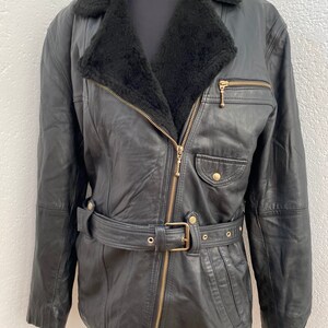 K&ouml;nnte beinhalten: Eine schwarze Lederjacke mit einem Kunstfellkragen und einem G&uuml;rtel. Die Jacke hat einen Rei&szlig;verschluss und goldfarbene Beschl&auml;ge.