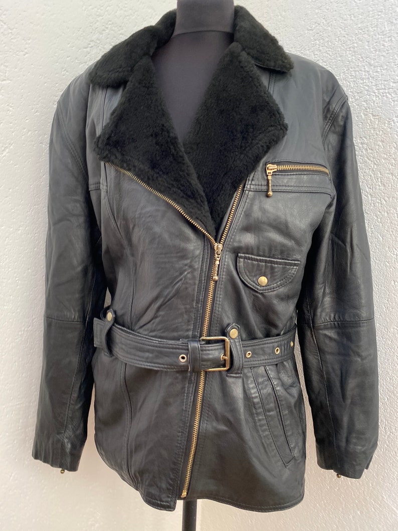 K&ouml;nnte beinhalten: Eine schwarze Lederjacke mit einem Kunstfellkragen und einem G&uuml;rtel. Die Jacke hat einen Rei&szlig;verschluss und goldfarbene Beschl&auml;ge.