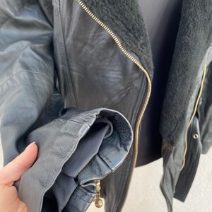 K&ouml;nnte beinhalten: Schwarze Lederjacke mit Rei&szlig;verschluss und Pelzkragen. Die Jacke hat eine Rei&szlig;verschlusstasche auf der linken Seite und einen goldenen Rei&szlig;verschluss.
